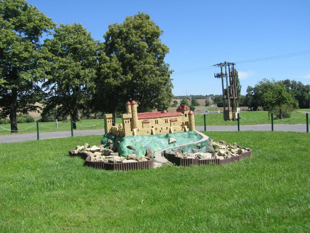 Minigolfplatz mit Rasenfläche, umzäunt mit Pfählen und Ketten, Bäumen, Strommasten mit Drähten und einer bewölkten Himmel im Hintergrund.