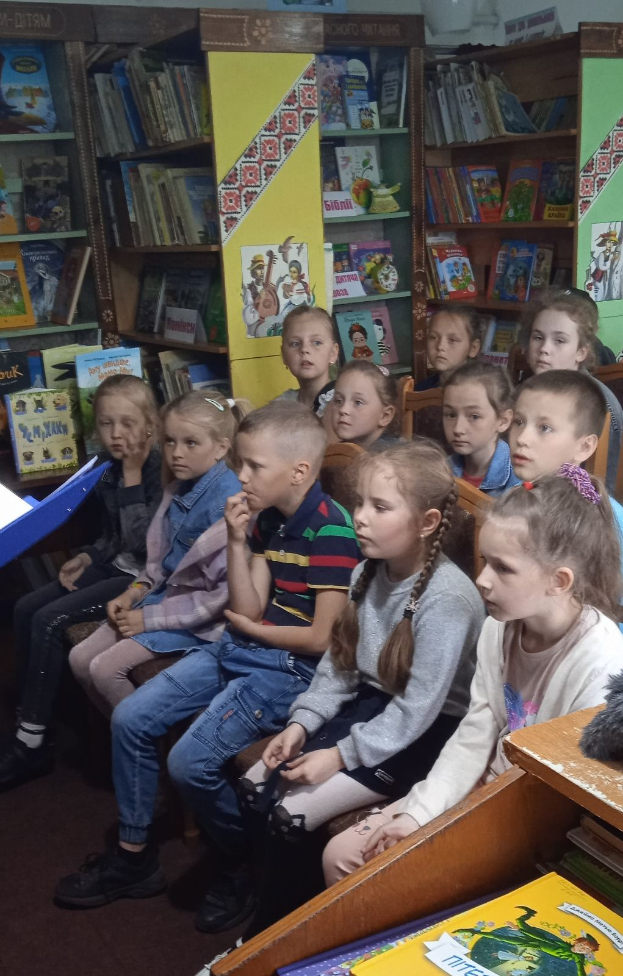 Eine Gruppe von Kindern sitzt in einer Bibliothek umgeben von Bücherregalen, vertieft in das Lesen von Büchern an Tischen.