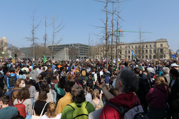 Große Menschenmenge mit Schildern und Taschen vor einem Gebäude mit Bäumen und einem klaren Himmel, was auf eine Klimaprotest Demonstration hinweist.