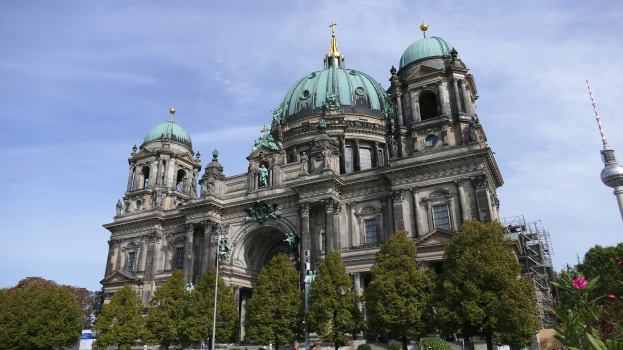 Großes, prunkvolles Berliner Dom in Berlin, Deutschland, mit architektonischen Details wie Säulen, Bögen und Statuen, vor einem bewölkten Himmel, mit Passanten, Bäumen und einem Turm im Vordergrund.