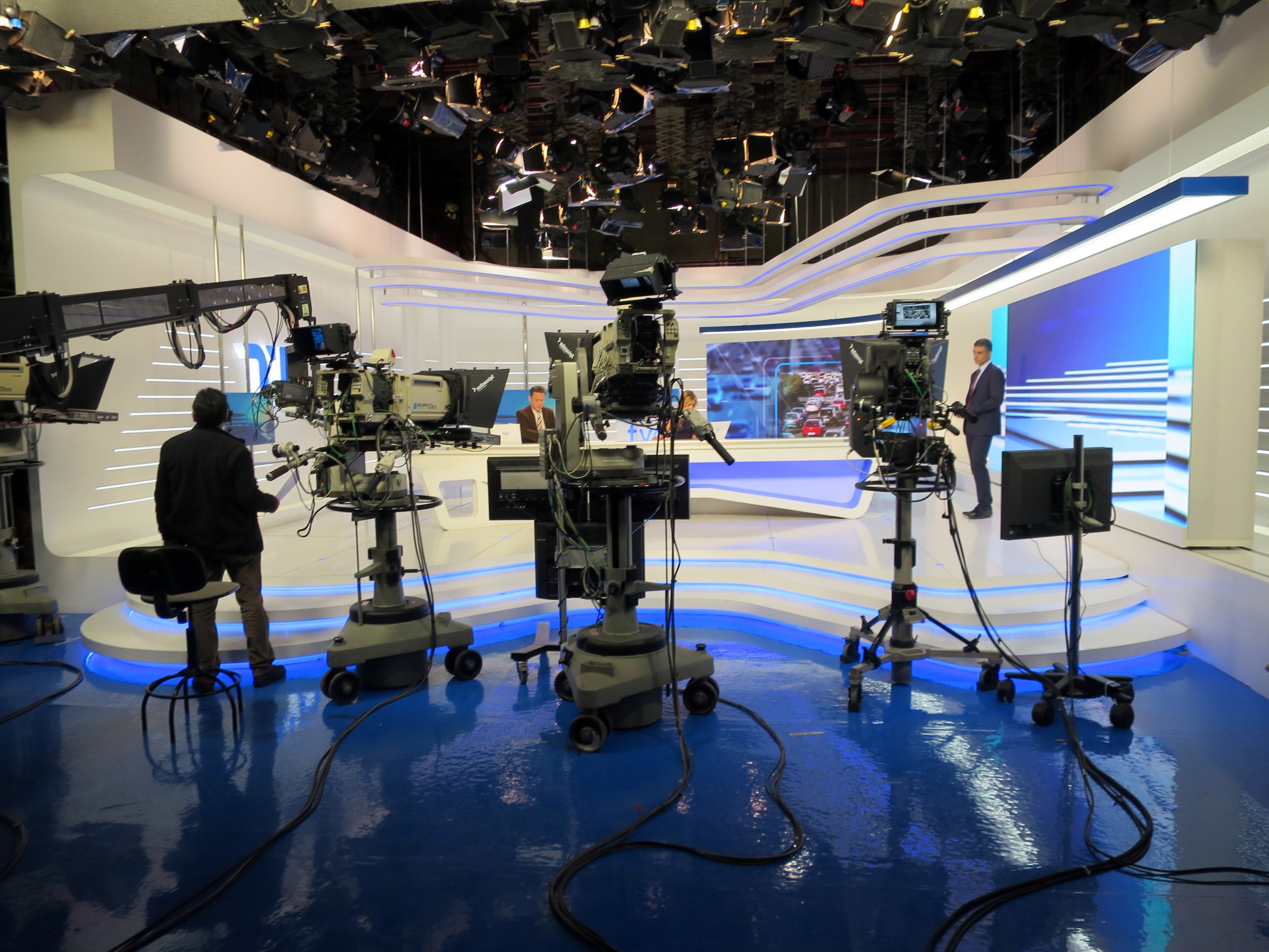 Ein TV-Studio mit mehreren Kameras auf dem Boden, einer sitzenden Person im Vordergrund und einigen stehenden Personen im Hintergrund, mit an der Decke montierten Lichtern für eine Livesendung.