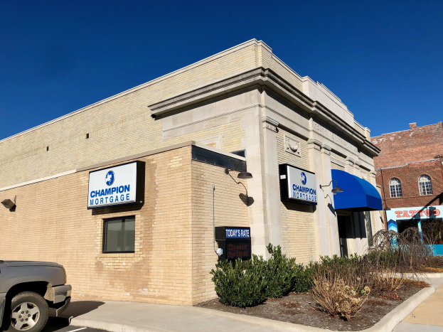 Ein zweistöckiges Gebäude mit vernagelten Fenstern, einem geparkten Auto, Topfpflanzen und Bäumen unter einem bewölkten Himmel, mit einem Schild, auf dem "Champion Mortgage, Inc." steht