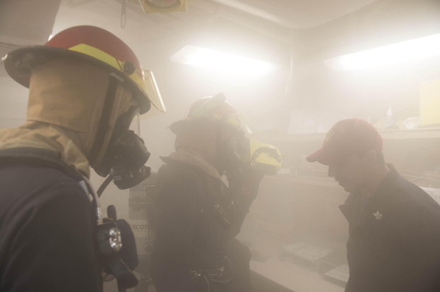 Feuerwehrleute in Helmen mit Rauch aus dem Mund, einer hält eine Kamera, während einer Übungssitzung mit Lichtern und Gegenständen im Hintergrund.