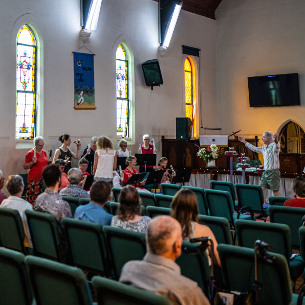 Eine Gruppe von Menschen sitzt auf Stühlen in einer Kirche, ein Mann steht vorne mit einem Mikrofon, umgeben von Musikinstrumenten, einem Tisch mit einer Blumenvase, einem Lautsprecher, einer Wand mit Text, Fenstern und Deckenleuchten.