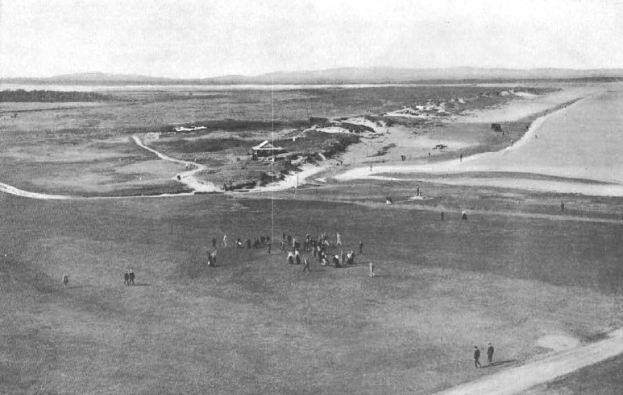 Schwarzes und weißes Foto von Golfern auf dem 18. Loch des Royal Birkham Golf Clubs, mit saftig grünem Rasen, verstreuten Häusern, sanften Hügeln und einem hellblauen Himmel im Hintergrund.