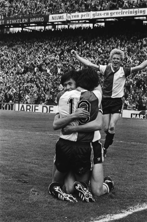 Drei Fußballspieler umarmen sich auf dem Boden nach einem Torerfolg, mit jubelnder Menge und schwenkenden Fahnen in einem schwarz-weißen Stadion.