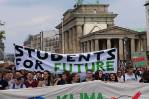 Gruppe von Schülern marschiert in Berlin mit einem leuchtend bunten "Students for Future"-Schild vorbei an Gebäuden, Bäumen und Himmel.