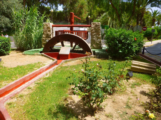Minigolfplatz in einem Park mit Grünflächen, einer Straße mit Fußgängern und Schildern sowie Bäumen vor einem blauen Himmel.