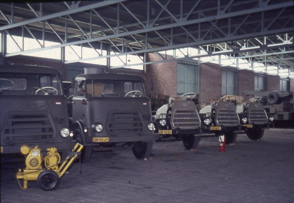 Eine Gruppe von Lastwagen, die in einem Gebäude mit einem sichtbaren Feuerlöscher, Metallstangen, Fenstern, Deckenleuchten und Rädern geparkt sind.