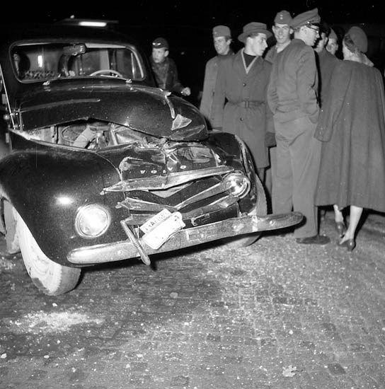 Schwarz-weißes Foto eines schwer beschädigten Autos am Straßenrand mit eingedellter Motorhaube und zersplitterten Fenstern, umgeben von Menschen, die es inspizieren.