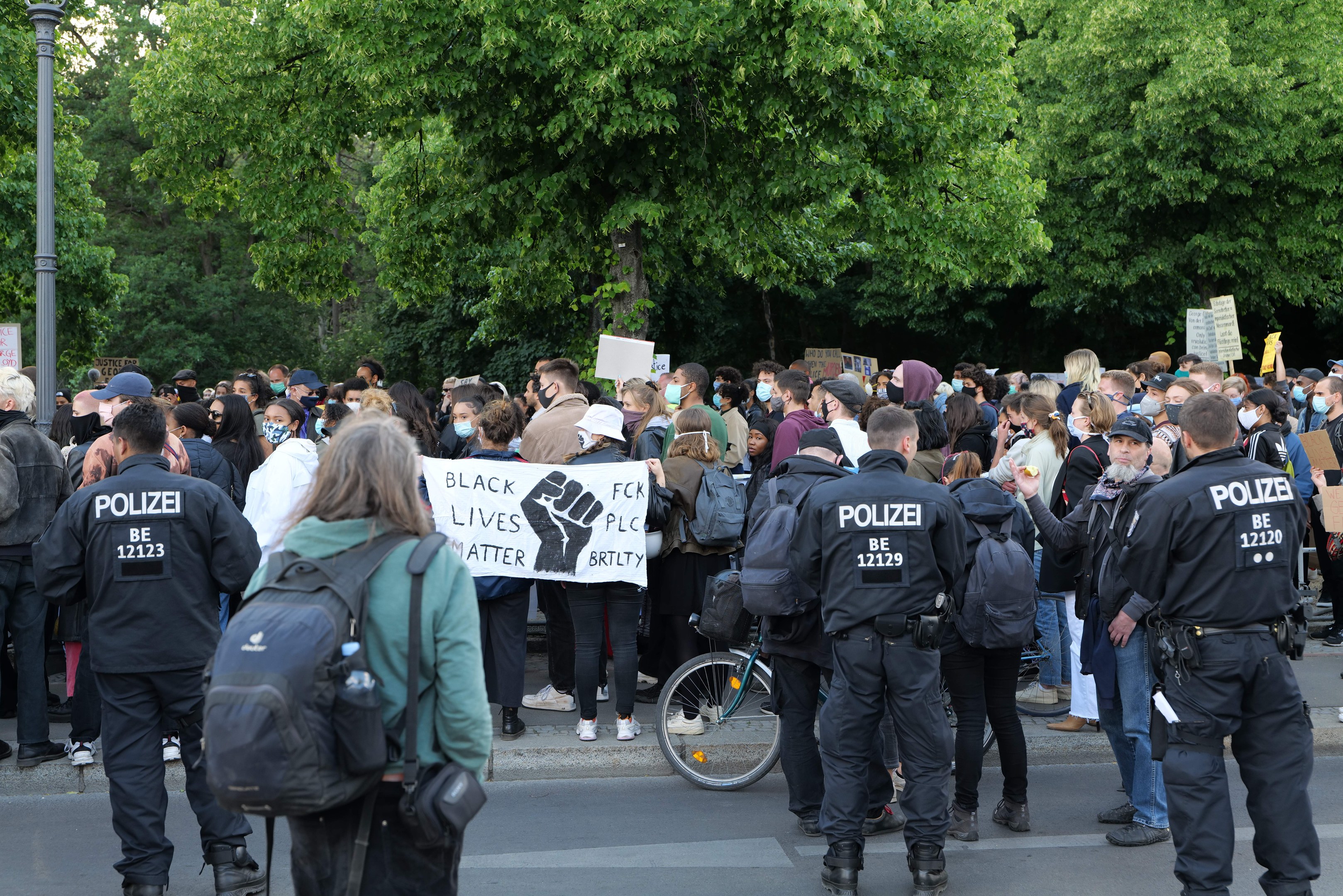 Eine große Gruppe von Menschen bei einer Black Lives Matter Demonstration in Berlin, einige halten Schilder, mit einem Fahrrad im Vordergrund und Bäumen und einem Pfahl im Hintergrund.