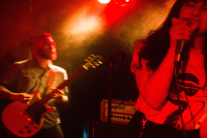 Eine Frau singt in ein Mikrofon auf der Bühne, während ein Mann Gitarre spielt und eine Deckenbeleuchtung den Hintergrund erhellt.