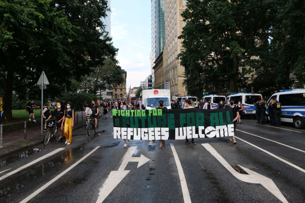Eine Gruppe von Menschen, die eine Straße entlanggehen und ein Banner mit der Aufschrift 'Flüchtlinge Willkommen' tragen, mit parkenden Fahrzeugen und Fahrradfahrern daneben, unter einem Himmel, der über Bäume und Gebäude sichtbar ist.