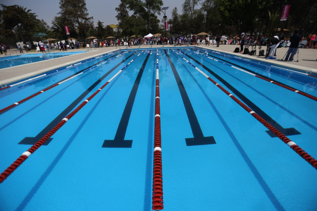 Großes Schwimmbad mit Bahnentrennern, umgeben von Menschen mit Taschen und Gegenständen, sowie Zelten, Transparenten, Stangen, einem Schild und einem bewölkten Himmel im Hintergrund.