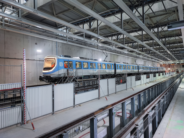 Blauer und weißer Zug fährt durch eine große Bahnhofshalle mit einer Plattform auf der rechten Seite, einer Wand im Hintergrund und einer Decke mit Stangen und Lampen, Teil eines neuen Zugsystems in Deutschland.