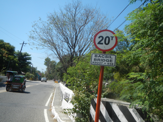 Eine Straße mit fahrenden Fahrzeugen, ein Tempolimit-Schild, eine Mittellinie, Bäume, Strommasten mit Kabeln und einen klaren blauen Himmel im Hintergrund.