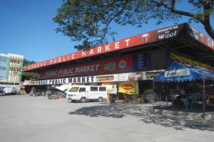 Öffentlicher Markt mit einem weißen Van vorne geparkt, umgeben von Gebäuden, Motorfahrzeugen, Menschen unter einem Zelt sitzend, Geländern, Bäumen und einem klaren blauen Himmel.