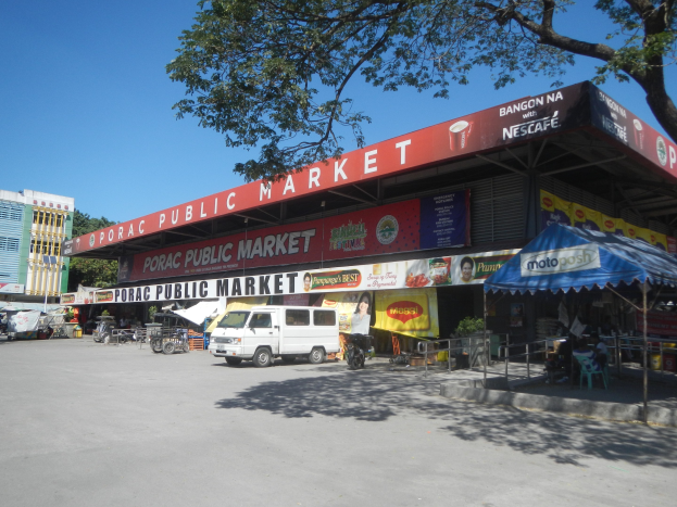 Öffentlicher Markt mit einem weißen Van vorne geparkt, umgeben von Gebäuden, Motorfahrzeugen, Menschen unter einem Zelt sitzend, Geländern, Bäumen und einem klaren blauen Himmel.