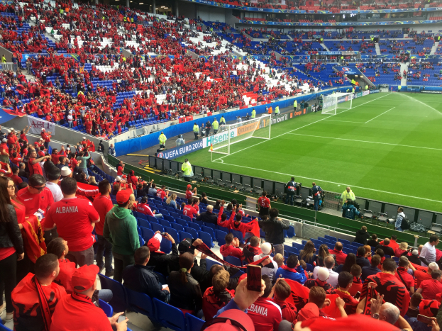 Ein volles Stadion mit Sitzplätzen und Stehplätzen, Fahnen, ein Tor und Deckenleuchten, während die Menge gespannt auf den Beginn eines Fußballspiels wartet.