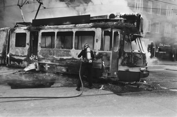 Schwarz-weißes Bild eines Mannes in einem Helm, der eine Pfeife h├Ąlt und neben einer ausgebrannten Stra├čenbahn auf einer Stra├če mit Rauch, Dr├Ąhten und einem Geb├Ąude im Hintergrund steht.