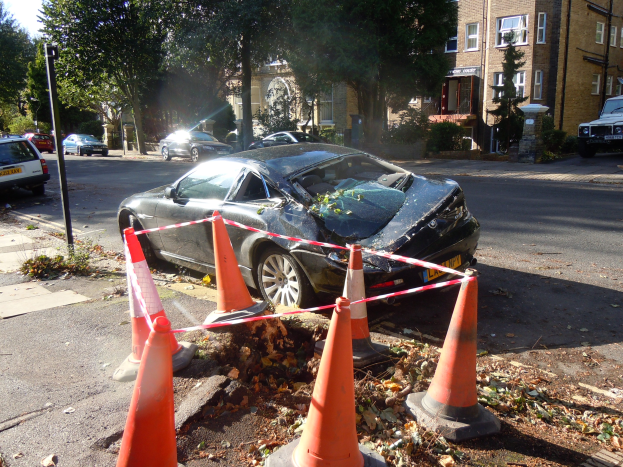 Ein beschädigtwagen am Straßenrand mit orangen Verkehrskegeln, Bäumen, Gebäuden und einem klaren blauen Himmel im Hintergrund.
