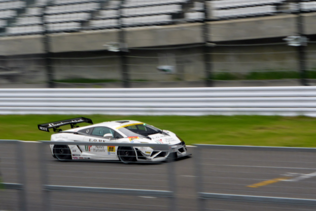 Weißer Sportwagen auf einer Rennstrecke, umgeben von einem Zaun und Gras, mit einem unscharfen Hintergrund, der hohe Geschwindigkeit andeutet.