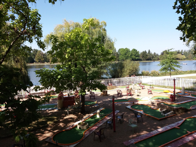 Minigolfplatz in einem Park mit grünem Gras, Bäumen und einem Zaun, umgeben von verstreuten Stühlen und Tischen, mit einem Gewässer und einem Boot im Hintergrund unter einem sichtbaren Himmel.