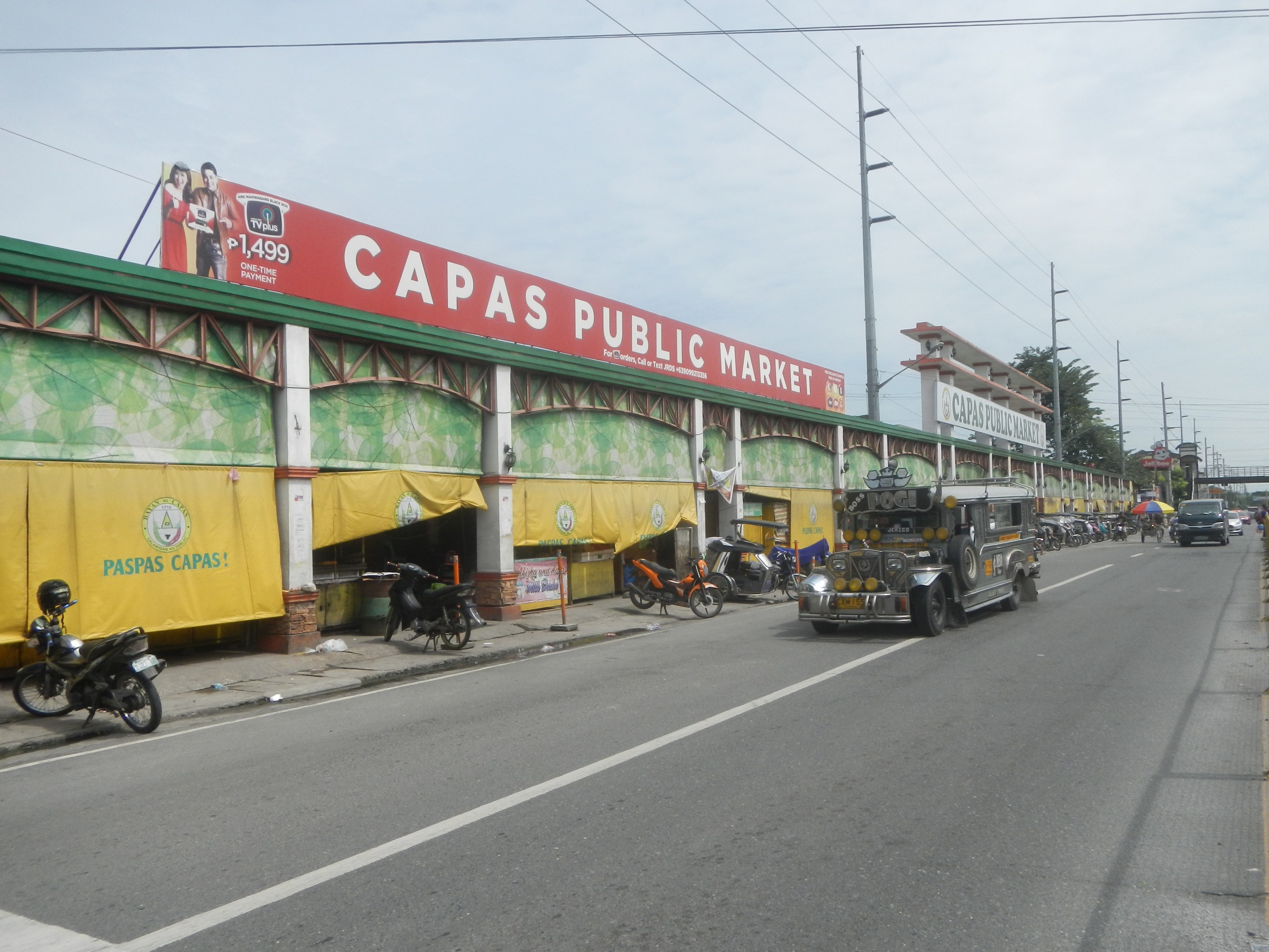 Eine belebte Straße mit Fahrzeugen, einem Gehweg, Strommästen, Gebäuden, Bäumen und einem bewölktem Himmel, mit einem Gebäude mit der Aufschrift "Capas Public Market" im Vordergrund.