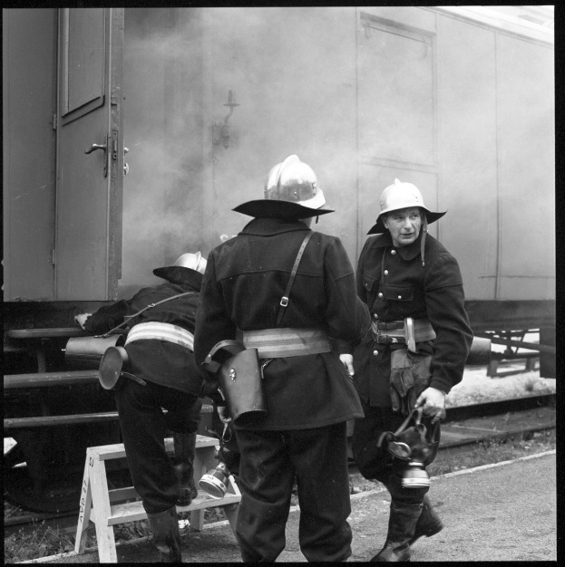 Feuerwehrmänner mit Helmen stehen auf einem Bahngleis, einer sitzt auf einem Hocker, während ein Zug mit qualmendem Rauch im Hintergrund in einem Schwarz-Weiß-Bild vorbeifährt.
