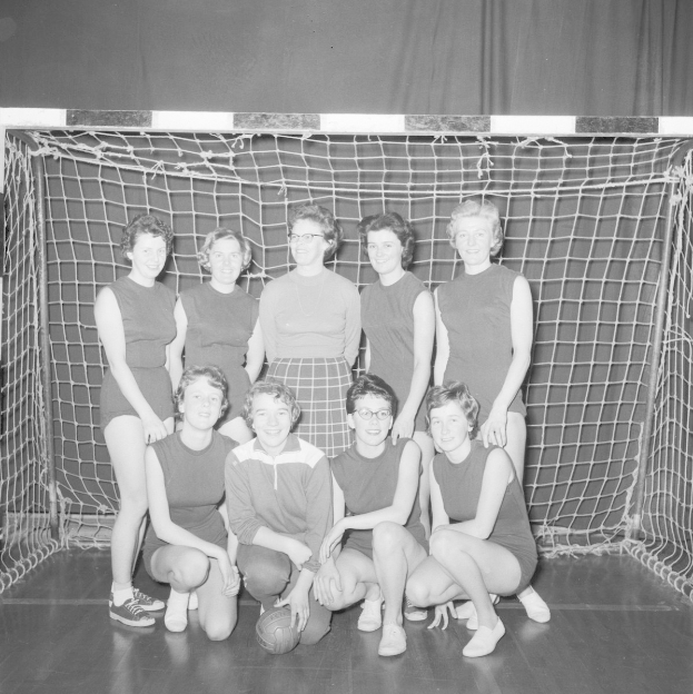 Schwarzes und weißes Foto einer Gruppe von Frauen, die vor einem Tor für ein Foto posieren, mit einem Ball auf dem Boden und einem Vorhang im Hintergrund.