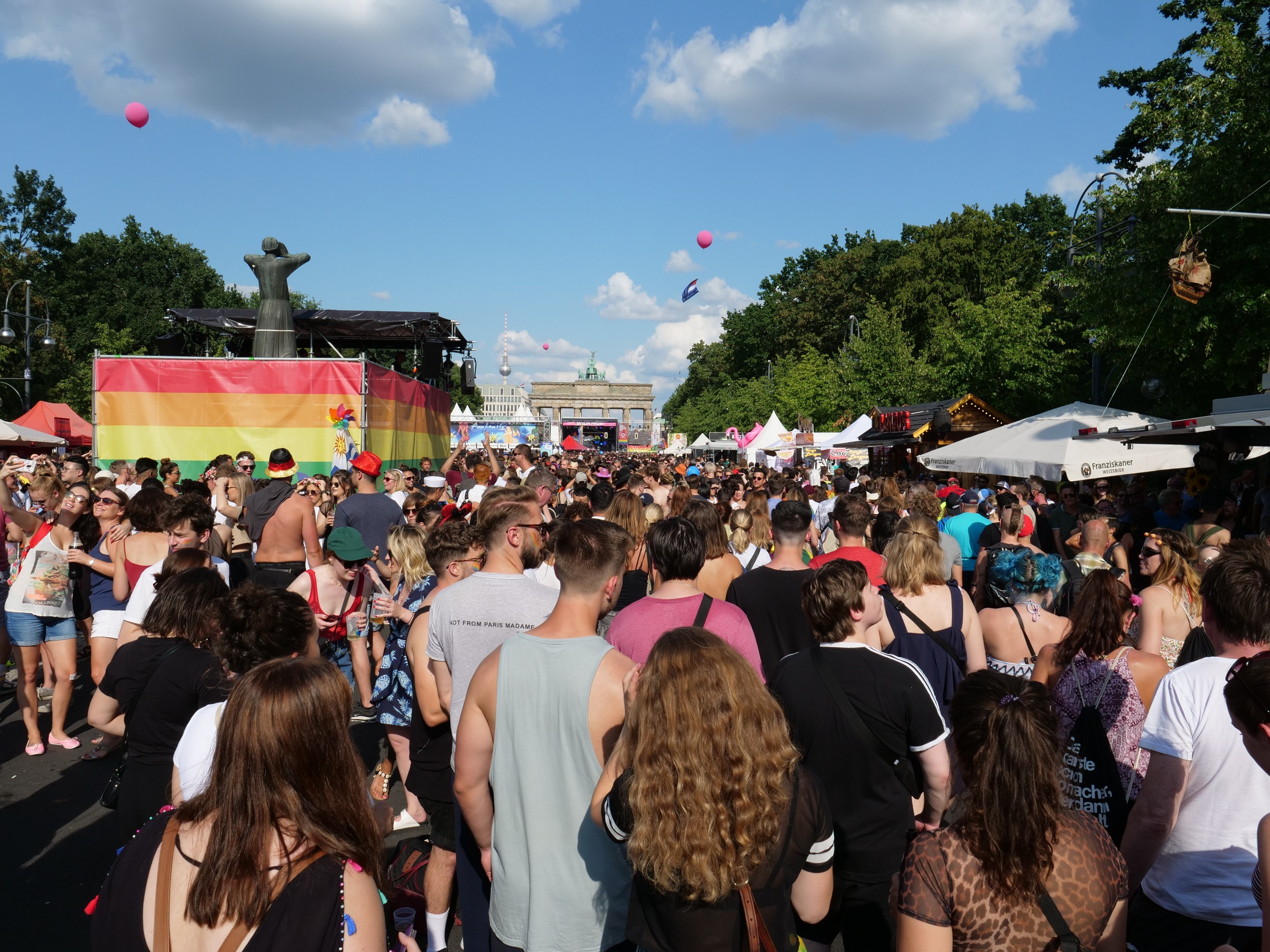 Eine große Menschenmenge geht eine Straße mit Zelten, Bäumen, Pfählen, Lichtern und einer Statue entlang während des Christopher Street Day Festivals in Berlin, mit Gebäuden und einem bewölkten Himmel mit Ballons im Hintergrund.