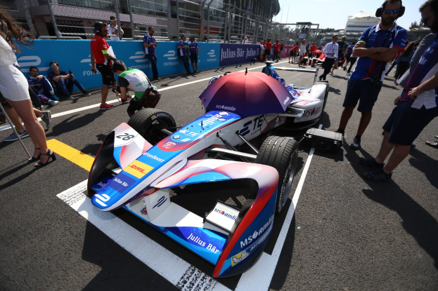 Formula E-Rennwagen mit weißer, blauer, roter und schwarzer Lackierung auf einer Rennstrecke geparkt, umgeben von Menschen, mit einem Schirm im Vordergrund und Rennstreckenabsperrungen, Gebäuden, Bäumen und einem klaren blauen Himmel im Hintergrund.