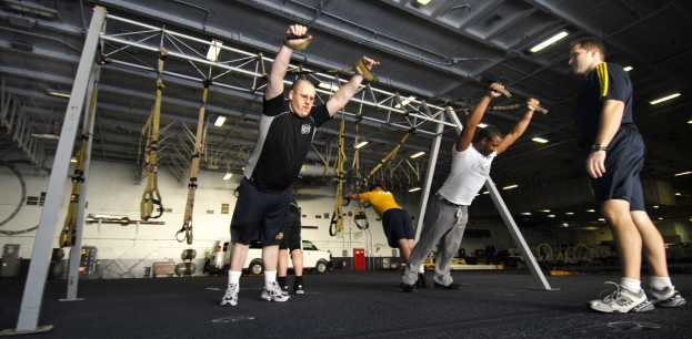 Mannergruppe beim Pull-up in einem Fitnessstudio mit Metallgeräten und Deckenbeleuchtung, mittig ein Mann mit Schuhen.
