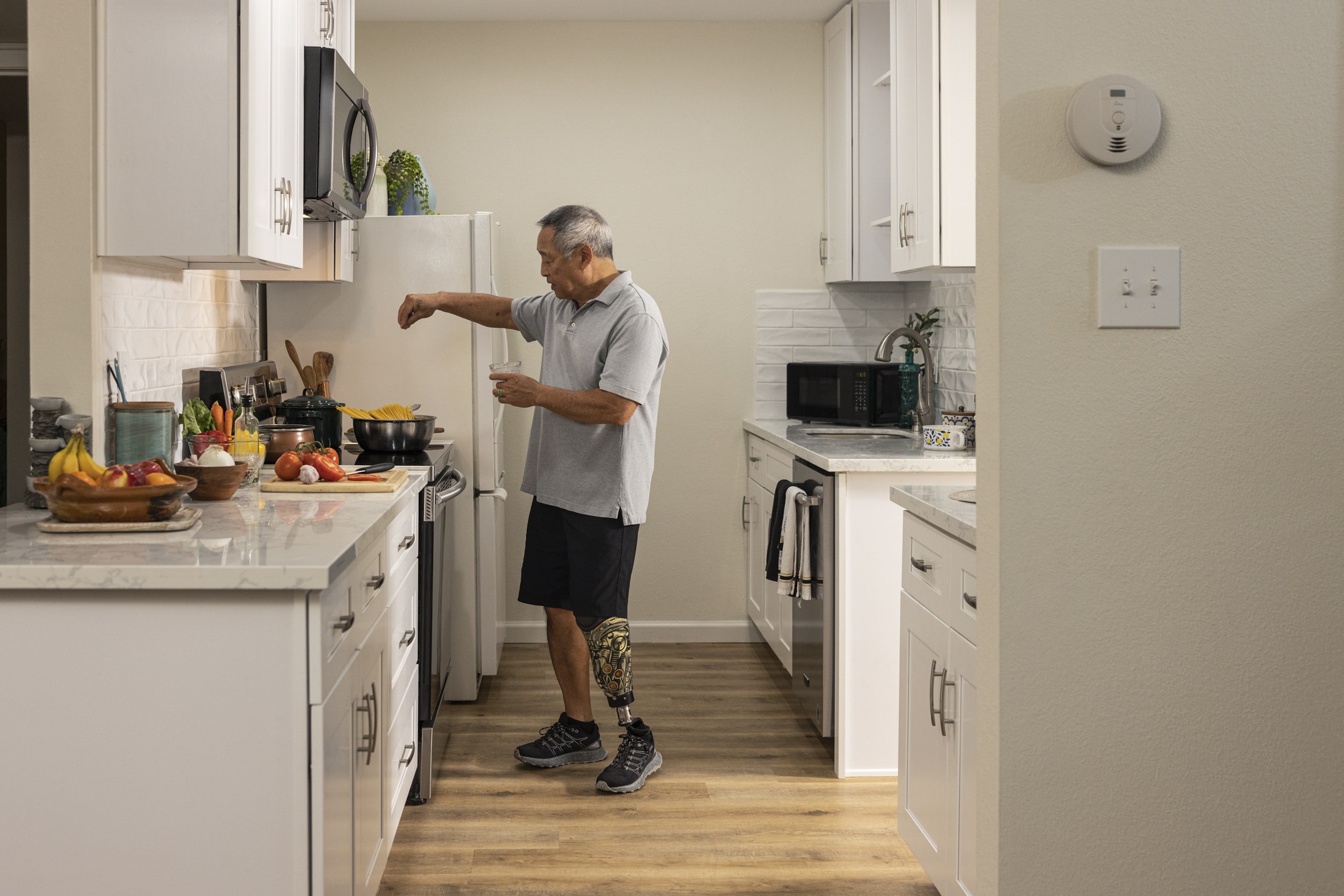 Ein Mann steht in einer Küche mit weißen Schränken und hält ein Glas in der Hand, während verschiedene Gegenstände wie Früchte, Gemüse, Schüsseln und Löffel auf einer Arbeitsplatte links von ihm liegen.