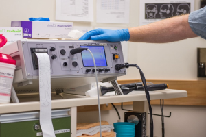 Eine Person in blauen Handschuhen arbeitet an einem medizinischen Gerät an einem unordentlichen Tisch in einem Krankenhauszimmer mit Papieren an der Wand.
