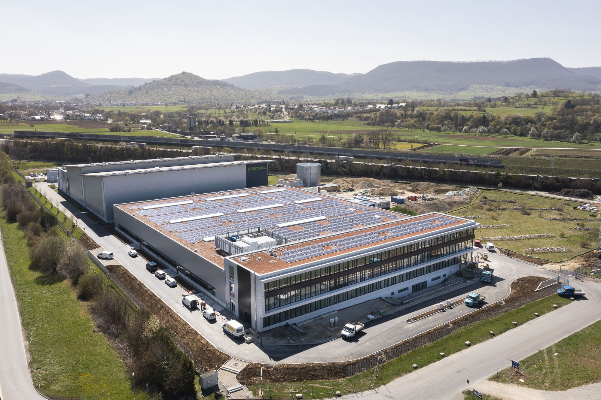 Luftaufnahme eines großen Gebäudes mit Solarmodulen auf dem Dach, umgeben von Fahrzeugen, Bäumen, Gras und Masten, vor einem Hügel- und klaren blauen Himmel, als Deutschlands erste solarbetriebene Fabrik bekannt.