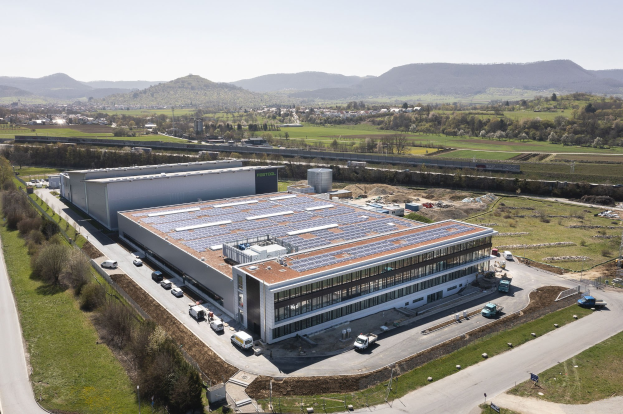 Luftaufnahme eines großen Gebäudes mit Solarmodulen auf dem Dach, umgeben von Fahrzeugen, Bäumen, Gras und Masten, vor einem Hügel- und klaren blauen Himmel, als Deutschlands erste solarbetriebene Fabrik bekannt.