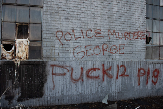 Graffiti an einem Gebäude mit der Aufschrift 'Polizei ermordet George Fuck 12 P19' mit Fenstern und verstreuten Gegenständen davor.