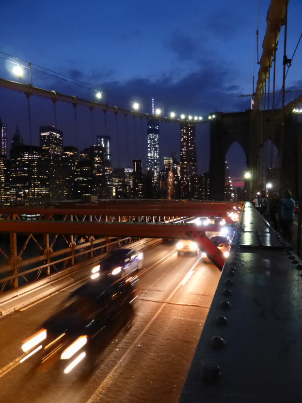 Ein Auto fährt über die Brooklyn Bridge bei Nacht, mit beleuchteten Gebäuden und Sternen im Hintergrund.