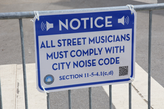 Anschlagtafel an einem Zaun mit einem Schild, auf dem "Anschlag: Alle Straßenmusiker müssen der Stadtlärmverordnung Folge leisten" steht, und eine Straße im Hintergrund.