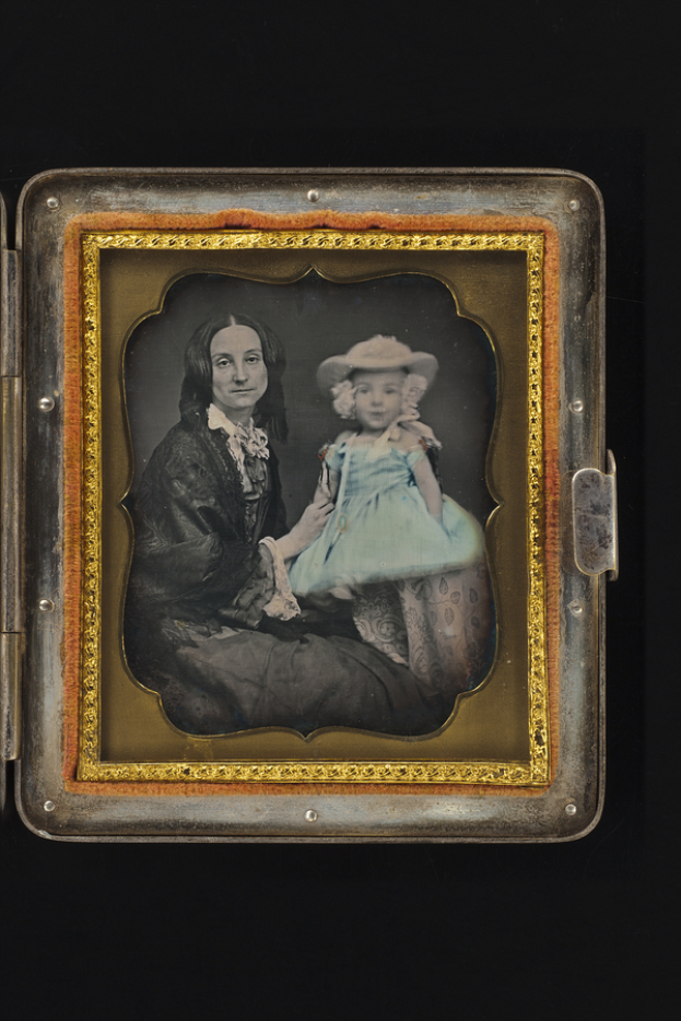 Ein altes Foto in einem dekorierten Rahmen zeigt eine Frau mit sanftem Ausdruck und ein Kind, das neugierig zu ihr hochschaut, beide mit gedämpften Farben.