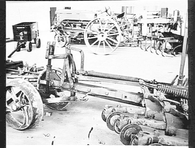 Schwarzes und weißes Foto eines Traktors in einer Fabrik, mit einer Egge im Vordergrund und einer Wand im Hintergrund, eingerahmt von einem Rahmen.