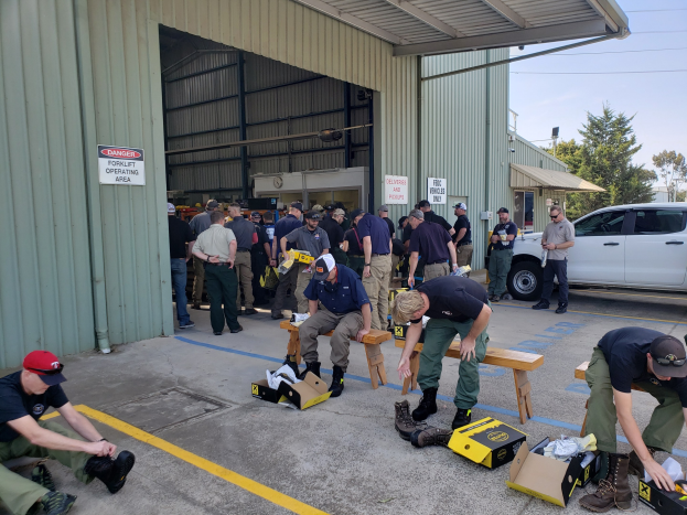 Eine Gruppe von Menschen bei einer Ausbildungssitzung der Nationalgarde, einige sitzen auf Bänken und andere stehen, mit verstreuten Kartons, Schuhen und Gegenständen vor einem Gebäude mit Fahrzeugen, Bäumen und Gebäuden im Hintergrund unter einem klaren blauen Himmel.