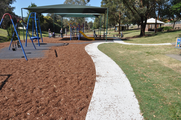 Ein Park mit einem Spielplatz mit Schaukeln und Rutschen, umgeben von grünem Gras, Bäumen, Bänken, Hütten und einem klaren blauen Himmel.