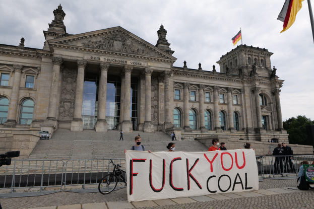 Protestierende mit einem "Fuck You Coal"-Schild vor dem Reichstaggebäude in Berlin, Deutschland, mit sichtbaren architektonischen Details, einem Fahrrad in der Nähe, Bäumen, einer Fahnenstange und einem bewölkten Himmel.