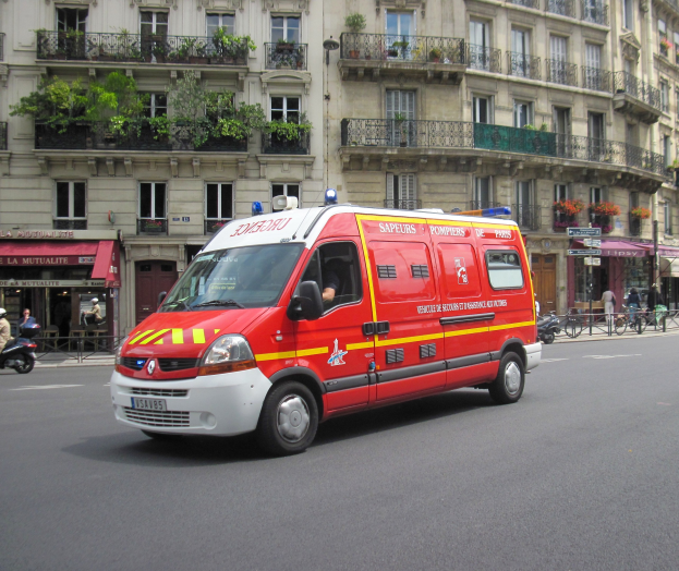 Ambulanz steht auf der Straßenseite vor einem Gebäude mit Fenstern, Geländern, Pflanzen und anderen Gegenständen, mit einigen Menschen und Fahrrädern im Hintergrund.
