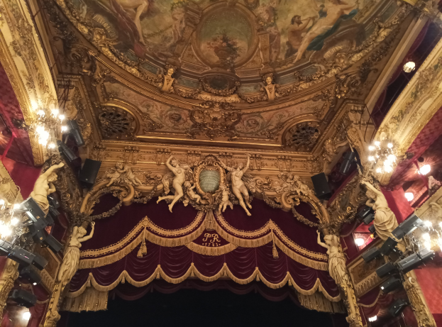 Theaterinnenraum mit roter Gardine, Kronleuchtern, Deckenplastiken, Lichtern und einem Gemälde, das eine Bühne und eine kleine Gruppe von Menschen am Boden zeigt.