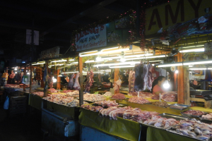 Eine nächtliche Szene im Schlachthof mit verschiedenen Fleischsorten, einigen Kunden und einer Waage auf der rechten Seite, mit beleuchteten Schildern im Hintergrund.
