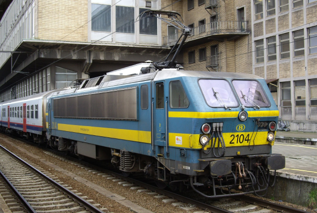 Blauer und gelber Elektrolokomotive fährt auf Schienen neben einem Glasgebäude mit einer Plattform und Menschen auf der rechten Seite.