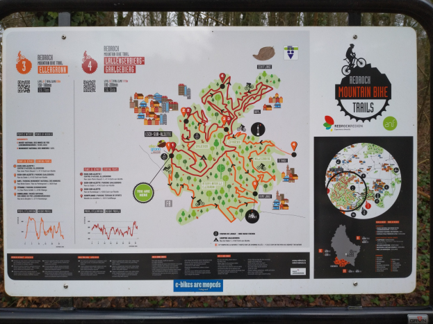 Ein Metallwegweiser in einem Waldgebiet mit einer Karte einer Mountainbike-Strecke mit Text und Illustrationen, vor einem klaren blauen Himmel.